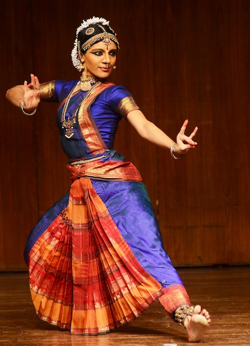 Aparna in bright purple traditional classical Indian dance garb. Her left foot is flexed and her fingers are splayed